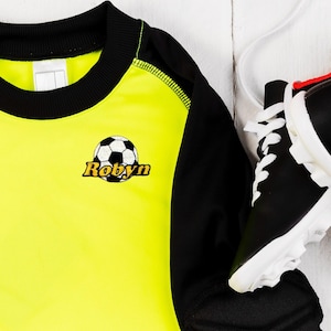 May include: Yellow and black soccer jersey with a black soccer ball and the name "Robyn" embroidered on the front.