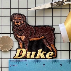 May include: Embroidered patch featuring a Rottweiler dog with the name "Duke" in gold thread. The dog is brown and black, with a black border. A quarter coin and a ruler are next to the patch.