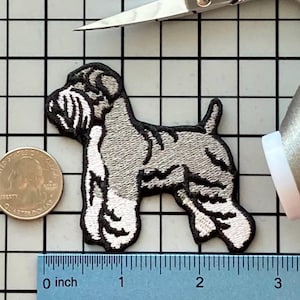Op de afbeelding: Geborduurde patch van een Schnauzer-hond in grijs, wit en zwart, met een zwarte rand. De patch ligt naast een munt, een schaar, een meetlint en een garenklos. De patch is ongeveer 7.6 cm lang.