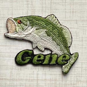 May include: An embroidered patch featuring a green and white largemouth bass fish with an open mouth. Below the fish, the name Gene is stitched in bold, dark green letters with a black outline, creating a detailed graphic design for apparel.