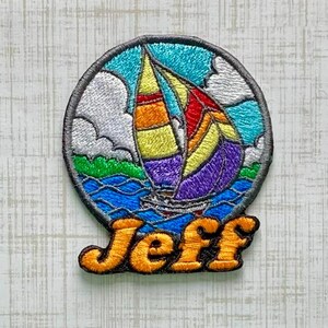 May include: A circular embroidered patch featuring a colorful sailboat with yellow, red, and purple sails on blue water under a sky with white clouds. The name Jeff is stitched in bold orange letters at the bottom of the circular design.