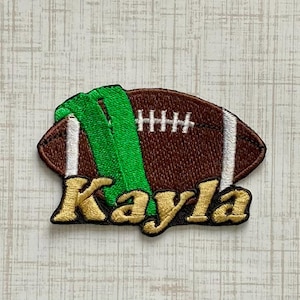 May include: An embroidered patch shaped like a brown football with white laces and a bright green stripe across the center. The name Kayla is stitched in gold thread with a black outline across the bottom portion of the football design.