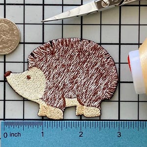 May include: Embroidered hedgehog patch with brown and cream colors. The patch is approximately 2 inches long. A quarter coin and a spool of thread are next to the patch. A pair of scissors is in the background.