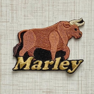 May include: An embroidered patch featuring a brown bull with cream-colored horns standing above the name Marley written in gold thread. The bull is set against a black background, and the entire design is outlined in a dark brown border.