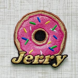 May include: An embroidered patch shaped like a donut with pink icing and colorful sprinkles. The center of the donut is black. Below the donut, the name Jerry is embroidered in gold thread with a black outline, featuring a decorative font style.