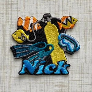 May include: An embroidered patch featuring a yellow scuba tank, blue swim fins, a blue diving mask, and two orange and white clownfish. The name Nick is stitched in large blue letters at the bottom against a black background with white outlines.