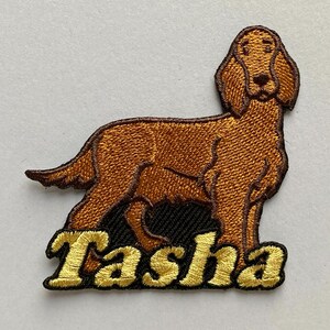 May include: An embroidered iron-on patch featuring a brown dog standing in profile with long ears. Below the dog, the name Tasha is stitched in bold, yellow, serif-style lettering with a black outline against a dark background.