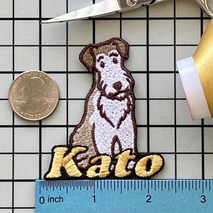May include: Embroidered patch featuring a seated dog with brown and white fur, and the name "Kato" in gold lettering. The patch measures approximately 2 inches in length. A quarter coin and a ruler are included for size reference.