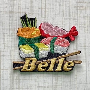 May include: An embroidered patch featuring a colourful sushi assortment with green, yellow, and pink elements, positioned above two gold-coloured chopsticks. The word Belle is stitched in gold thread across the chopsticks in a bold, serif font.