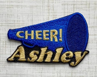 Könnte beinhalten: Ein blauer, bestickter Aufnäher in Form eines Megafons mit dem goldenen Text CHEER! und dem Namen Ashley in goldener Schreibschrift darunter. Der Aufnäher hat einen dunkelblauen Rand und eine strukturierte Oberfläche mit sichtbaren Nähten.