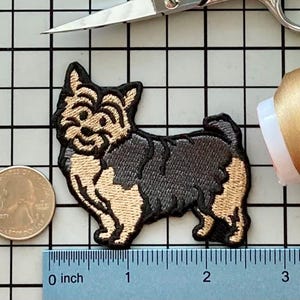 May include: Embroidered Yorkshire Terrier dog patch in black, tan, and cream. The patch is on a grid with a ruler showing inches. A quarter coin and scissors are also in the image.