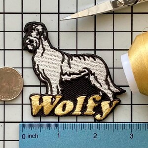 Pode incluir: Patch bordado com um cão e o nome "Wolfy" em letras douradas. O cão é branco e preto. O patch tem aproximadamente 7,6 cm de comprimento. Uma régua é visível.