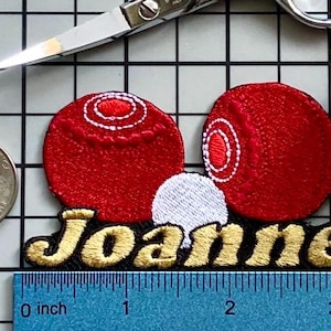 Puede incluir: Parche bordado con dos círculos rojos, detalles blancos y un círculo blanco entre ellos. El nombre "Joanne" está bordado con hilo dorado sobre un fondo negro. Una moneda y una regla están al lado del parche.