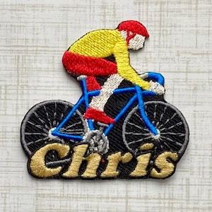 May include: An embroidered patch featuring a cyclist wearing a yellow jersey, red shorts, and a red helmet riding a blue bicycle. The word Chris is stitched in gold thread across the bottom of the patch over a black background.