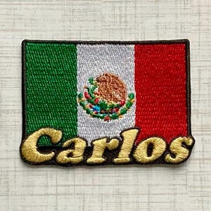 May include: A rectangular embroidered patch featuring the flag of Mexico with its central emblem of an eagle on a cactus. The name Carlos is stitched in gold-colored thread across the bottom of the patch, which has a dark brown border.