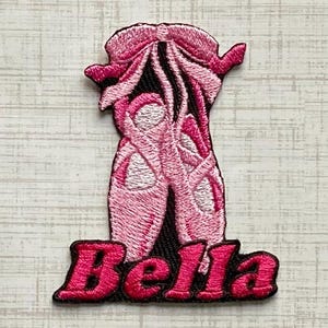 May include: An embroidered patch featuring a pair of light pink ballet slippers with dark pink ribbons tied in a bow at the top. The name Bella is stitched in bold, dark pink capital letters across the bottom of the patch on a black background.
