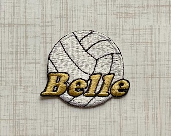 Könnte beinhalten: Ein runder Stickaufnäher, der einen weißen Volleyball mit schwarzen Nahtlinien zeigt. Das Wort Belle ist in der Mitte des Volleyballs in einer fetten, goldfarbenen Schrift mit einem dicken schwarzen Rand und einer strukturierten Optik gestickt.