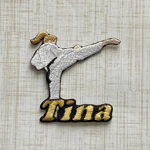 May include: An embroidered patch featuring a white martial arts uniform silhouette in a high kick pose with gold hair and the name Tina stitched in gold thread with a black outline at the base of the figure on a textured off-white background.