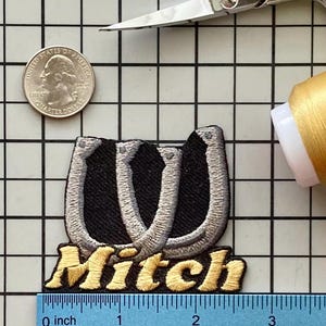 May include: Embroidered patch with a black horseshoe design and the name "Mitch" in gold lettering. The patch is approximately 2 inches wide. A quarter coin and scissors are also in the image.