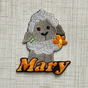 May include: An embroidered patch featuring a gray and white lamb holding a small orange flower. Below the lamb, the name Mary is stitched in bold orange letters with a dark outline. The design is rendered in detailed thread work on a fabric base.