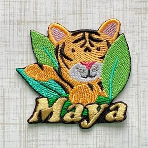 May include: An embroidered patch featuring a tiger face with orange fur, black stripes, and a white muzzle, framed by bright green leaves. The name Maya is stitched in gold thread with a dark brown outline at the bottom of the patch design.