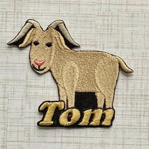 May include: An embroidered patch featuring a tan goat with dark gray horns and a pink nose. The goat is standing above the name Tom, which is stitched in gold-colored thread with a dark brown outline, set against a solid black background.