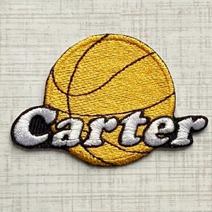May include: A circular embroidered patch featuring a yellow basketball with black lines. The name Carter is stitched across the center in white thread with a black outline, creating a layered effect over the basketball graphic design.