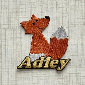 May include: An embroidered patch featuring a stylized orange fox with a white-tipped tail and white chest fur. The fox is positioned above the name Adley, which is stitched in gold-colored thread with a black outline on a dark background.
