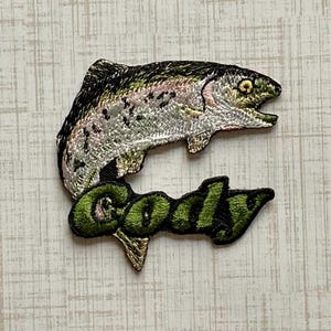May include: An embroidered patch featuring a silver and green fish leaping above the name Cody stitched in bold, textured green thread. The fish has detailed scales, a dark dorsal fin, and a visible eye, with black outlines defining the entire shape.