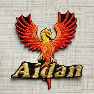 May include: An embroidered patch featuring a stylized phoenix with vibrant orange and red wings and a yellow head. The phoenix is positioned above the name Aidan, which is stitched in bold, gold-colored lettering with a black outline.