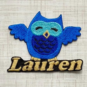 May include: An embroidered patch featuring a bright blue owl with a light blue face and a gold beak. The owl has dark blue wing details and a scalloped chest pattern. Below the owl, the name Lauren is stitched in gold thread on a black base.
