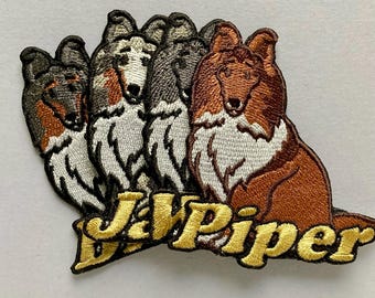 May include: An embroidered patch featuring four collie dogs in shades of brown, white, and grey. The dogs are arranged in a row, and the text "J&Piper" is stitched in bold, yellow, outlined letters across the bottom of the patch.