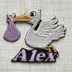 May include: An embroidered patch featuring a white stork with a gold beak carrying a purple bundle. Below the stork, the name Alex is stitched in purple letters on a black background. The design uses white, gold, purple, and black thread.
