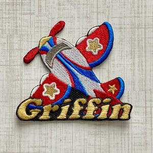 May include: An embroidered patch featuring a stylized airplane in red, white, and blue with gold star accents on the wings. The name Griffin is stitched in gold thread with a black outline at the base of the airplane design on a white background.