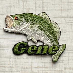 May include: An embroidered iron-on patch featuring a green and white largemouth bass fish leaping upward. Below the fish, the name Gene is stitched in bold, dark green letters with a black outline, creating a cohesive graphic design element.