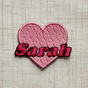 May include: A heart-shaped embroidered patch featuring a light pink textured background with a cable knit pattern. The name Sarah is stitched across the center in bold, dark pink thread with a black outline, creating a clear and distinct design.