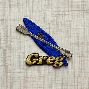 May include: An embroidered patch featuring a bright blue kayak positioned diagonally with a silver-grey paddle resting across it. Below the kayak, the name Greg is stitched in gold thread with a black outline, creating a distinct contrast.