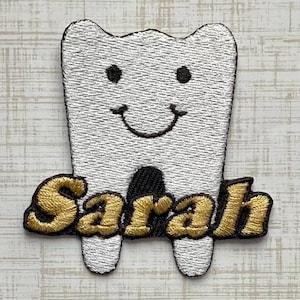May include: A white embroidered patch shaped like a smiling tooth with two black eyes and a black mouth. The name Sarah is stitched in gold thread with a black outline across the bottom of the tooth, overlapping the two roots of the tooth shape.