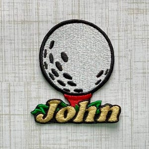 May include: An embroidered patch featuring a white golf ball with black dimples resting on a red tee. Below the tee, the name John is stitched in gold thread with a dark outline, accented by small green leaves on the left and right sides.