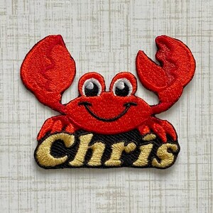 May include: An embroidered patch featuring a bright red crab with large claws and white eyes. Below the crab, the name Chris is stitched in gold thread against a black background. The design is detailed with dense red and gold embroidery stitching.