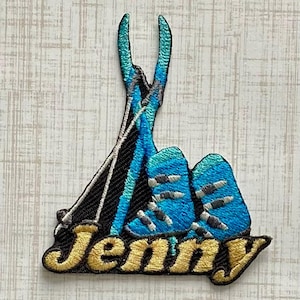 May include: An embroidered patch featuring a pair of bright blue ski boots and two crossed ski poles against a black background. The name Jenny is stitched in gold thread across the bottom in a cursive font with a black outline and shadow.
