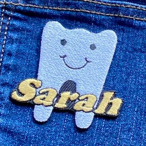 Puede incluir: Un parche bordado azul claro con forma de diente sonriente con el nombre "Sarah" en letras doradas. El parche está sobre un fondo de mezclilla azul.