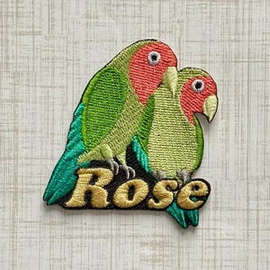 May include: An embroidered patch featuring two lovebirds with bright green bodies and coral-pink faces. The birds are perched above the word Rose, which is stitched in gold-coloured thread with a black outline against a dark background.