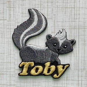 May include: An embroidered patch featuring a stylized skunk with dark gray fur and white stripes on its back and tail. The skunk is positioned above the name Toby, which is stitched in a bold, gold-colored font with a dark outline.