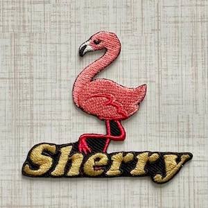 May include: An embroidered patch featuring a pink flamingo standing on top of the name Sherry, which is stitched in gold thread with a black outline. The flamingo has a white and black beak and is depicted in a side profile view with its neck curved.