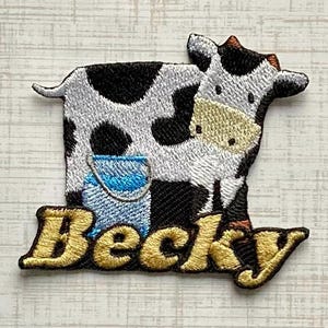 May include: An embroidered patch featuring a black and white cow standing next to a light blue bucket. The name Becky is stitched in gold thread across the bottom of the patch in a cursive font, positioned below the cow and the bucket.