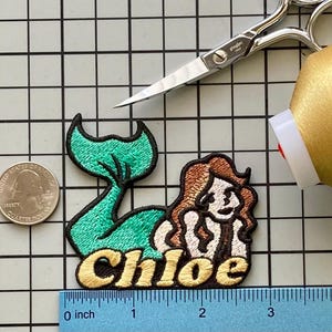 May include: Embroidered patch of a mermaid with a teal tail and brown hair, with the name "Chloe" in gold lettering. A quarter coin and a ruler are next to the patch. Scissors and a spool of gold thread are also in the image.