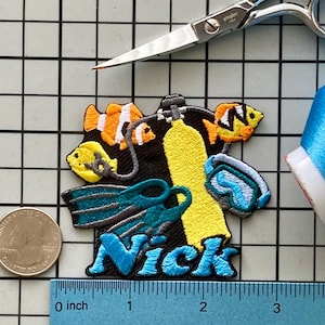 Puede incluir: Parche bordado con un diseño de buceo y el nombre "Nick" en azul. El diseño incluye un tanque de aire amarillo, aletas azules y peces de colores. Se incluye una moneda y una regla para la escala.