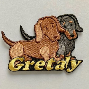 May include: An embroidered patch featuring two dachshund dogs, one light brown and one black and tan, standing side by side. Below the dogs, the word Gretaly is stitched in bold, yellow, cursive lettering against a dark background.