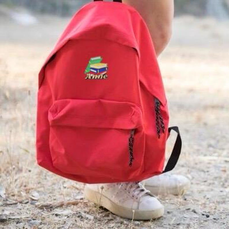 May include: Red backpack with a black zipper and a small embroidered patch of a stack of books with the word "Annie" underneath.